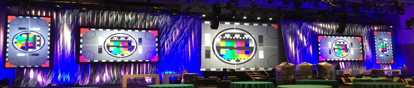 Event stage framed by pleated blue-and-purple uplit drapery, featuring five large LED video screens—two tall side screens, two medium screens, and one central oversized screen—all displaying standardized color-bar and grayscale test patterns for AV calibration, with round green-cloth tables set in front